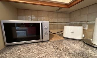 A modern kitchen with a microwave and a toaster on the countertop. The background consists of bright tiles and wooden cabinets.