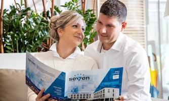 A couple is sitting together on a couch looking at a brochure. They are smiling at each other and appear relaxed.