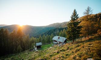 A picturesque mountain landscape at sunset with a wooden house and many trees. The gentle hills and the warm colors of the sky give the scene a tranquil atmosphere.