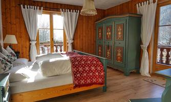 A cozy bedroom with wooden paneling and large windows. A stylish bed and a decorated wardrobe give the room a rustic charm.