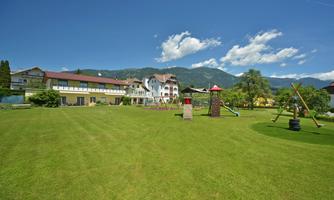 A spacious garden with a playground and a hotel building in the background. The landscape is surrounded by green meadows and beautiful mountains.
