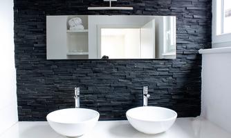 A modern bathroom with two white sinks and a large mirror. The wall is clad with black stone tiles.