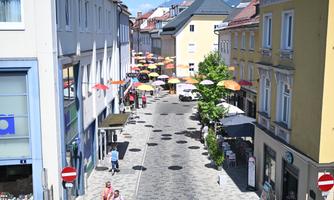 A bustling street with colorful umbrellas and shops. The sun is shining, and the atmosphere is welcoming.