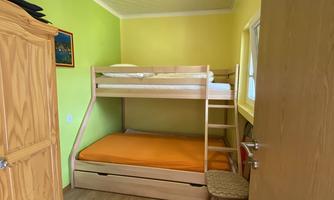 A small room with a bunk bed and an additional bed in orange. The walls are painted light green, and there is a wooden wardrobe.