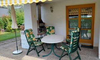 A cozy outdoor area with a round table and chairs. The space is equipped with a sunshade and a grill, surrounded by a well-kept garden.