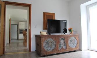 A cozy room with a beautifully decorated wardrobe and a television. In the background, there is an open kitchen and a window with plenty of daylight.