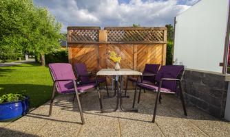 A cozy outdoor area with purple chairs and a small table. In the background, a green meadow and a fence are visible.