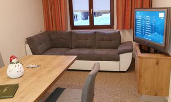 A cozy living space with a gray sofa and a table. A television is on a wooden piece of furniture next to a large window with curtains.