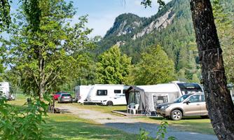 A beautiful campsite with caravans and cars, surrounded by trees and mountains. The landscape is green and inviting.