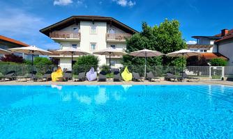A clear pool with loungers and sun umbrellas surrounded by green plants. In the background, several buildings can be seen under a blue sky.