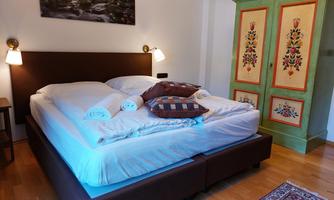 A cozy bedroom with a large bed and decorative pillows. The wardrobe has a colorful floral painting and the walls are light.