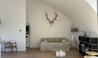 A bright living room with a sofa and a large window. On the wall hangs a deer antler, and there is a modern table as well as a floor lamp.