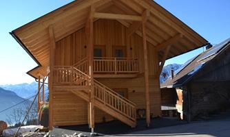 A modern wooden house with a large balcony and a staircase. In the background, mountains and a clear sky can be seen.