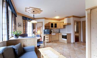 A bright kitchen with wooden furniture and a cozy living area.  
Large windows let in plenty of light and there are plants for decoration.