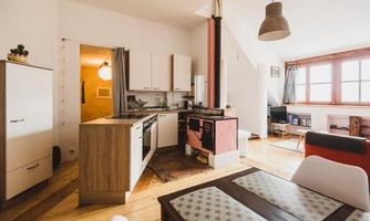 A modern kitchen with wooden flooring and white cabinets. There is a dining table and large windows for plenty of light.