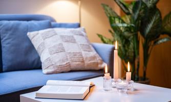 A cozy living room with a blue sofa and a decorative pillow. On the table, there are candles and an open book.