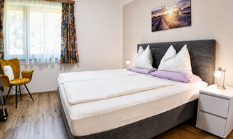 A cozy bedroom with a large box spring bed and bright pillows. A sliding wardrobe, a comfortable chair, and a window with curtains complete the room.