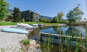 A calm pond with boats on the shore and lush vegetation. In the background, a modern building and a clear blue sky can be seen.