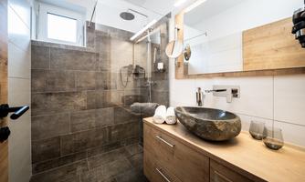A modern bathroom with a large shower and natural wood-colored accents. The decor is simple and stylish, featuring a round stone sink.
