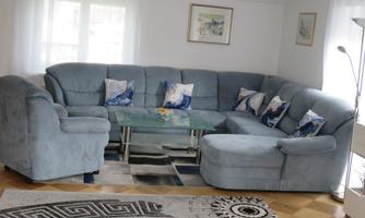 A comfortable blue sofa with cushions in various shades is arranged around a glass table. The room is bright and cheerful, with a rug and windows that let in daylight.