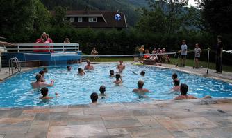 A swimming pool full of cheerful people playing in the water. In the background, trees and a residential building can be seen.