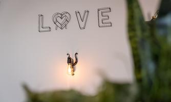 A wall with the word "LOVE" in wire design and a warm wall lamp. In the foreground, blurred plant elements are visible.