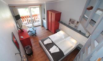 A modern bedroom with a double bed and wooden floors. Large windows offer a view of a balcony and contribute to a bright atmosphere.