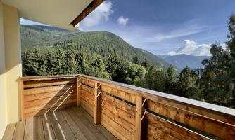 A balcony with a wooden railing and a view of green mountains. The sky is clear and blue with a few clouds.