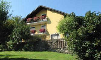A yellow house with balconies adorned with colorful flowers. Surrounded by green trees and a well-kept garden.