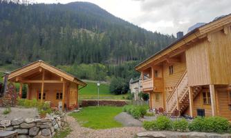 A picturesque place with two wooden houses and green meadows. In the background, forested mountains stretch under a partly cloudy sky.