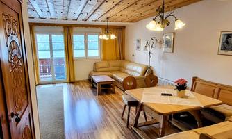 A cozy living room with wooden ceilings and large windows. It features a sofa, a table, and a corner bench that create an inviting atmosphere.