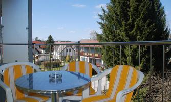 A balcony with a round table and chairs in yellow and white design. In the background, houses and trees can be seen against a clear sky.