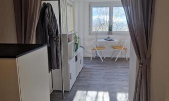 A bright room with white walls and a large window. In the foreground, there is a setup for a cozy sitting area with two chairs and a small table.