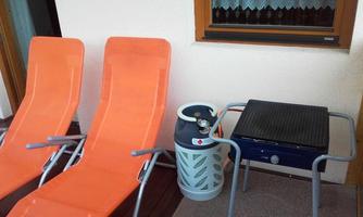 Two orange loungers stand next to a grill outdoors. In the background, a window with curtains can be seen.