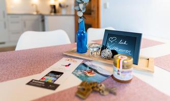 A beautifully set table with a blue vase, a welcome message, and various items like postcards and keys. In the background, a modern kitchen can be seen.