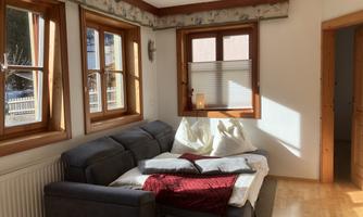 A cozy living room with a sofa adorned with a blanket and pillows. Bright windows allow plenty of daylight to enter.