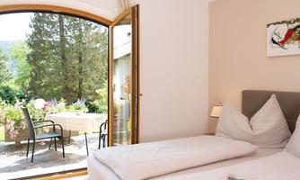 A bright bedroom with a large window that overlooks a beautiful garden view. Outside, there are chairs and a table on the terrace.