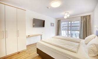 A modern bedroom with a double bed and a television on the wall. Large windows provide plenty of daylight and a view of a balcony.