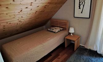 A cozy bedroom with a single bed and a nightstand. The floor is wooden, and a window curtain lets soft light in.