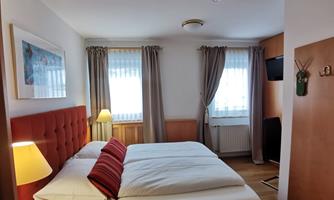 Cozy, harmonious bedroom with two beds and a red headboard. The room has large windows with curtains and a pleasant lighting.