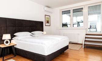 A bright bedroom with a large bed and a nightstand. The windows offer a view of a balcony and let in plenty of daylight.