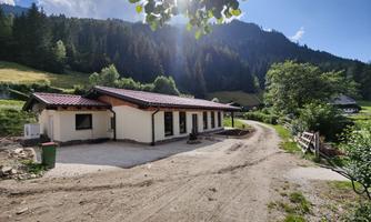A modern house in a rural setting with a view of the mountains. The sun is shining and there is plenty of green around.