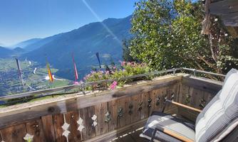 A balcony with a view of the valley and the surrounding mountains. Flowers adorn the railing and there is a radiant blue sky visible.