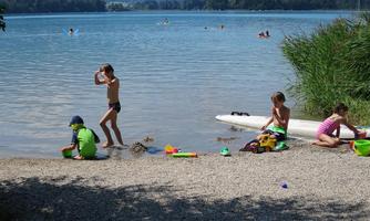 Children play at the shore of a lake and enjoy the summer day. In the background, there are more bathers and a beautiful landscape to be seen.