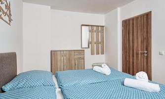 A bright room with two beds and blue bed sheets. On the wall there is a wooden shelf and a door.