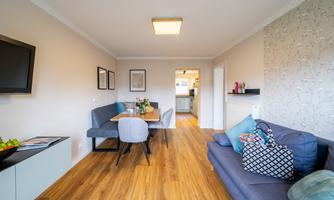 A modern living room with a sofa and a dining table. The room has wooden flooring and a bright, friendly atmosphere.