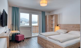A modern room with two beds and a wooden shelf. Large windows offer a view of the mountains and plenty of daylight.