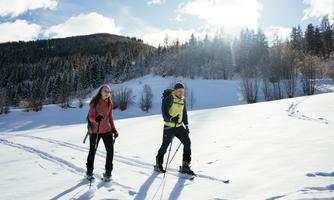 Two people are hiking on snow in a winter landscape. They are wearing warm clothing and enjoying the sunny atmosphere.