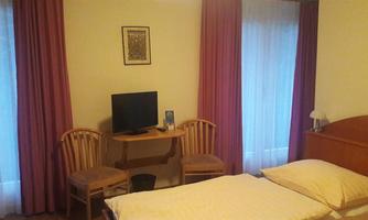 A simple room with a bed, two chairs, and a small table. A television is on the table, and light curtains let in daylight.