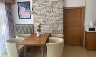 A cozy dining area with a wooden table and four bright chairs. In the background, a stone wall and a door can be seen.